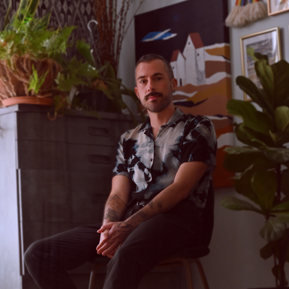 photo of Nico Stuart. He sits on a chair, surrounded by plants and art.